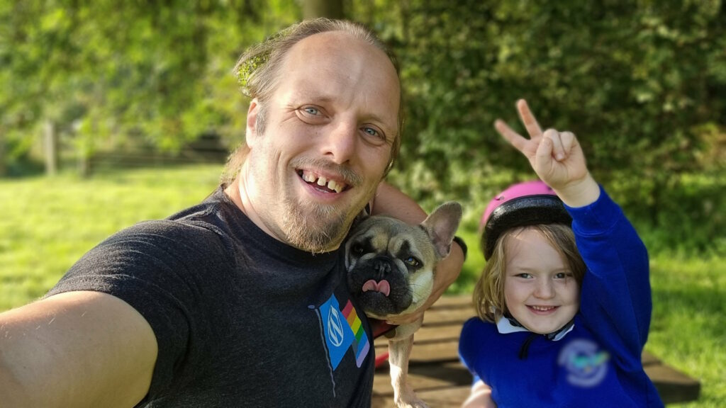 Dan, wearing a WordPress Pride "rainbow flag on black" t-shirt, sits on a park bench alongside a French Bulldog (with her tongue sticking out) and a young boy (throwing a peace sign, wearing a pink cycle helmet and a blue school uniform).