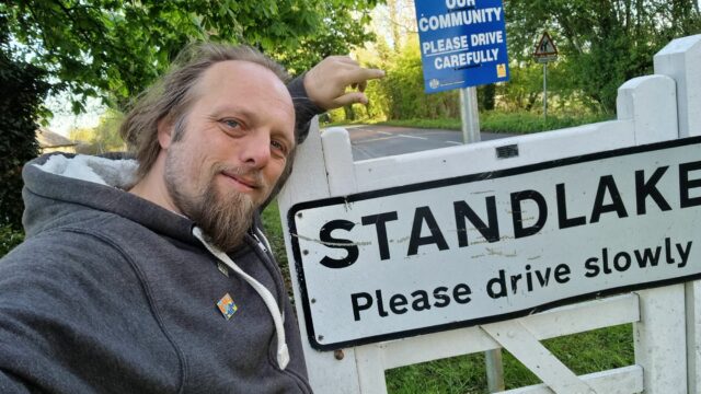 Dan, wearing a grey hoodie, stands by a sign welcoming visitors to the village of Standlake.