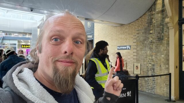 Dan, a queue of tourists behind him, points over his shoulder beyond them to a sign on a brick wall, "Platform 9¾".