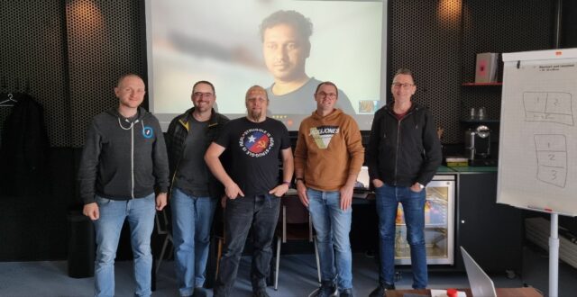 Dan stands with four other men in front of a projector screen with a fifth man on it.