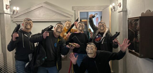 A group of people dressed as robbers (with masks covering their faces and automatic weapons in their hands) pose menacingly in the lobby of a vault.